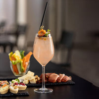 Copa transparente con cóctel rosa decorado con rodaja de naranja enrollada, hoja verde y bayas, sobre mesa negra con aperitivos y