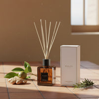 Difusor de ambiente marrón transparente con varillas, caja blanca con texto CULTI Milano, raíz de jengibre, hojas verdes y rama aromática sobre