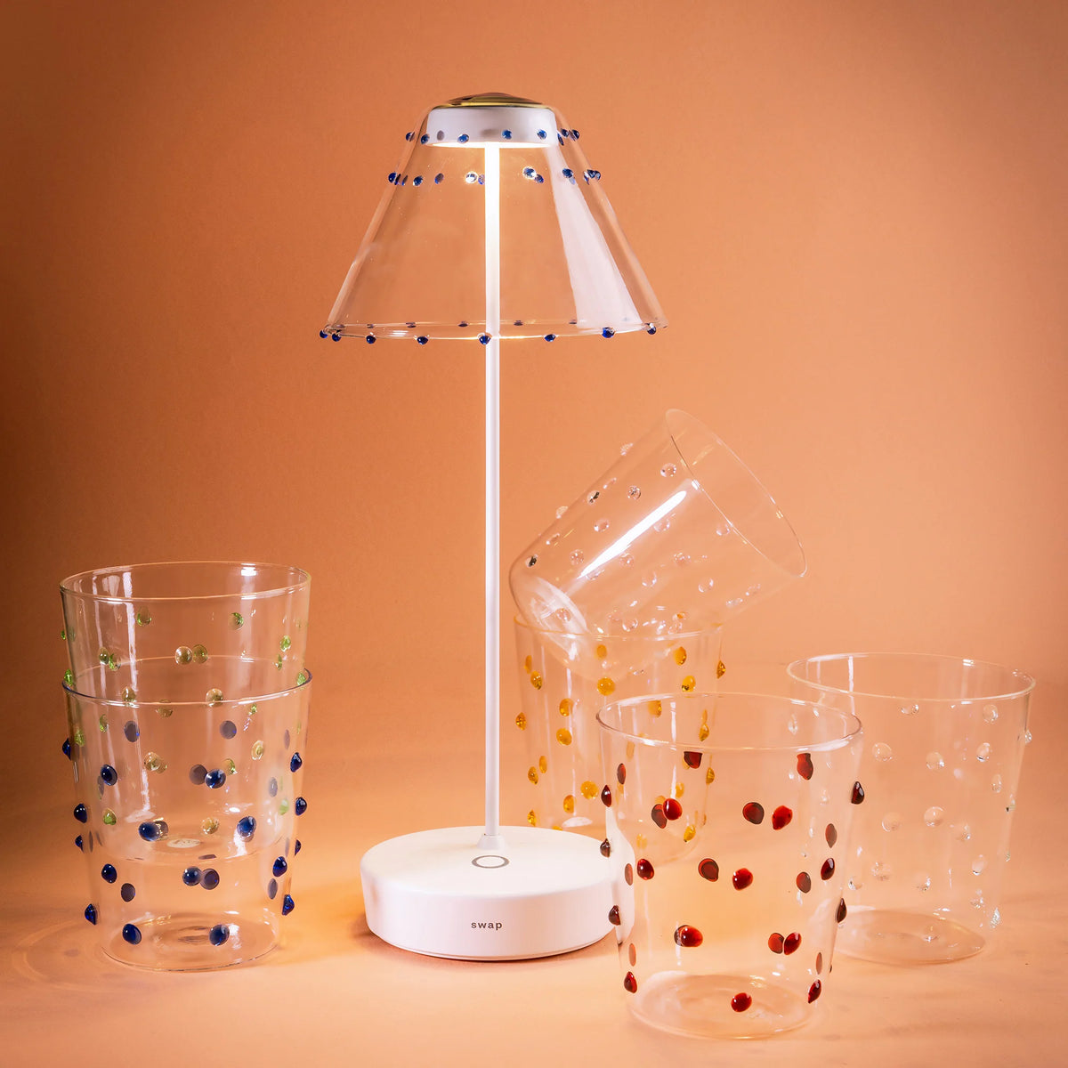 Lampada da tavolo bianca con paralume trasparente e decori blu, circondata da cinque bicchieri trasparenti con puntini colorati, sfondo arancione.