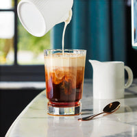 A coffee glass with poured milk, brown and cream shades, on a marble table with a teaspoon and white carafe.