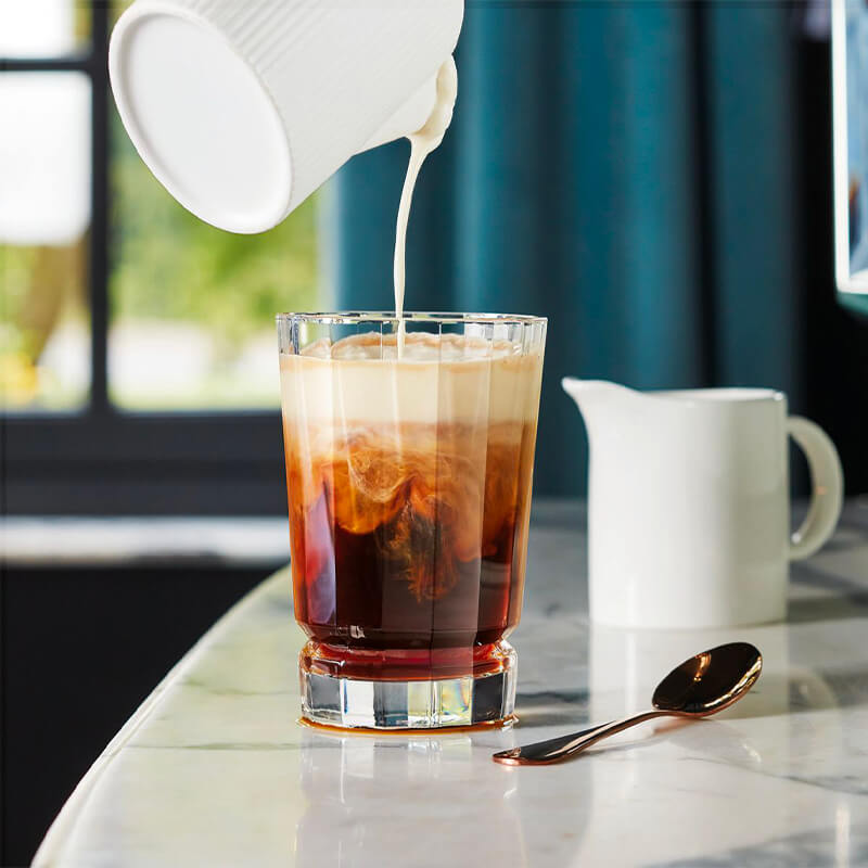 A coffee glass with poured milk, brown and cream shades, on a marble table with a teaspoon and white carafe.