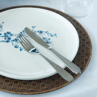 Piatti con decorazione floreale blu e posate d'acciaio con manico decorato su tovaglia bianca e sottopiatto marrone intrecciato.