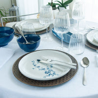 Tavola apparecchiata con piatti bianchi decorati con fiori azzurri, bicchieri trasparenti rigati, ciotole blu e posate metalliche su