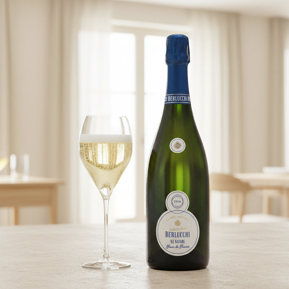 Botella de champagne Berlucchi 2016 junto a una copa con espumoso dorado, sobre una mesa con fondo luminoso y decoración sencilla.