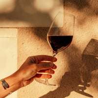 Main avec bracelet tenant un calice en verre transparent avec vin rouge contre un mur beige éclairé par une lumière chaude