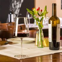 Bicchiere di vetro trasparente con vino rosso su tovaglietta bianca di tavolo di legno, bottiglia di vino, vasetto con fiore e piatto con posate.