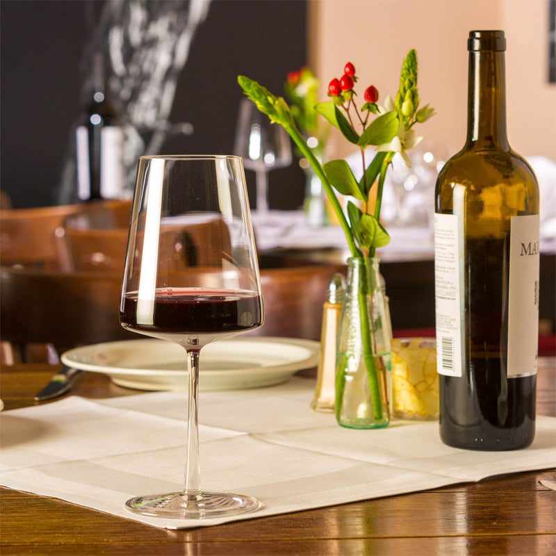 Bicchiere di vetro trasparente con vino rosso su tovaglietta bianca di tavolo di legno, bottiglia di vino, vasetto con fiore e piatto con posate.
