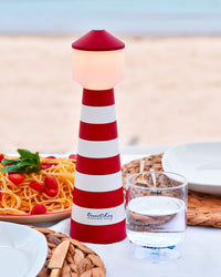 Rood-wit gestreepte vuurtoren tafellamp met bord spaghetti, waterglas en bestek op wit tafellaken.