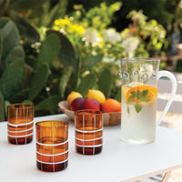 Trois verres en verre ambré avec décorations blanches et carafe transparente avec eau, tranches d'orange et menthe sur table blanche extérieure.