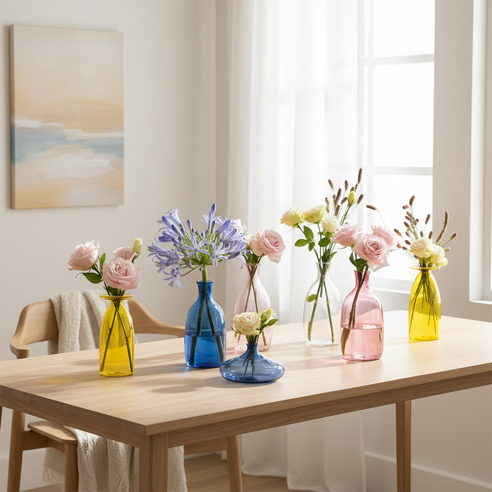 Set di sette vasi di vetro colorati (giallo, blu, rosa, trasparente) con fiori freschi su tavolo di legno chiaro in ambiente illuminato da finestra.