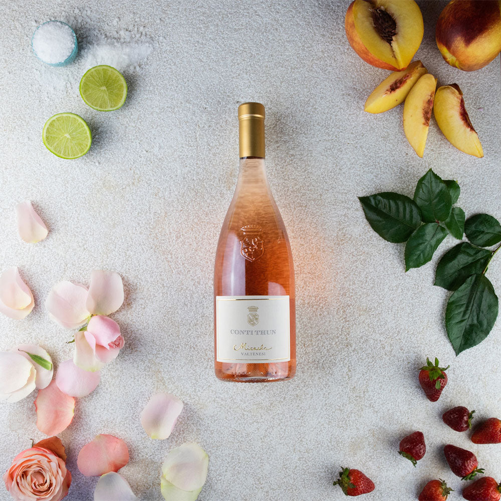 Botella de vino rosado Conti Thun Micasa con flores rosas, lima, melocotones, fresas y hojas verdes sobre fondo claro.