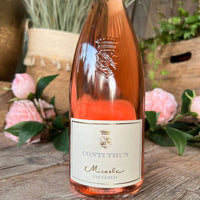 Botella de vino rosado Conti Thun con etiqueta blanca e inscripciones doradas, sobre mesa de madera con rosas rosas y plantas verdes al fondo
