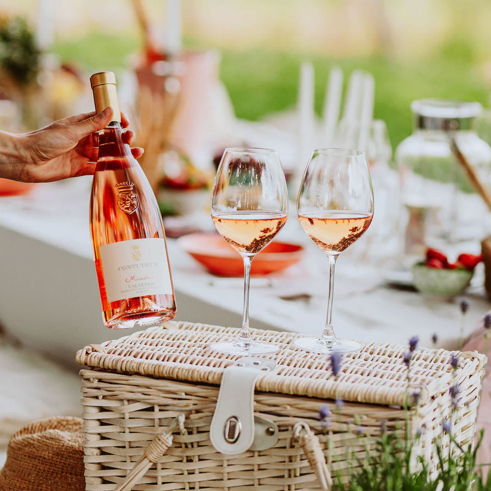 Botella de vino rosado CONTE VITUS y dos copas llenas en cesta de picnic de mimbre en ambiente exterior luminoso.