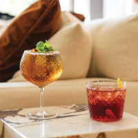 Deux verres transparents à surface facettée, contenant des boissons jaune ambré et rouge avec glace et garniture, sur table en marbre.