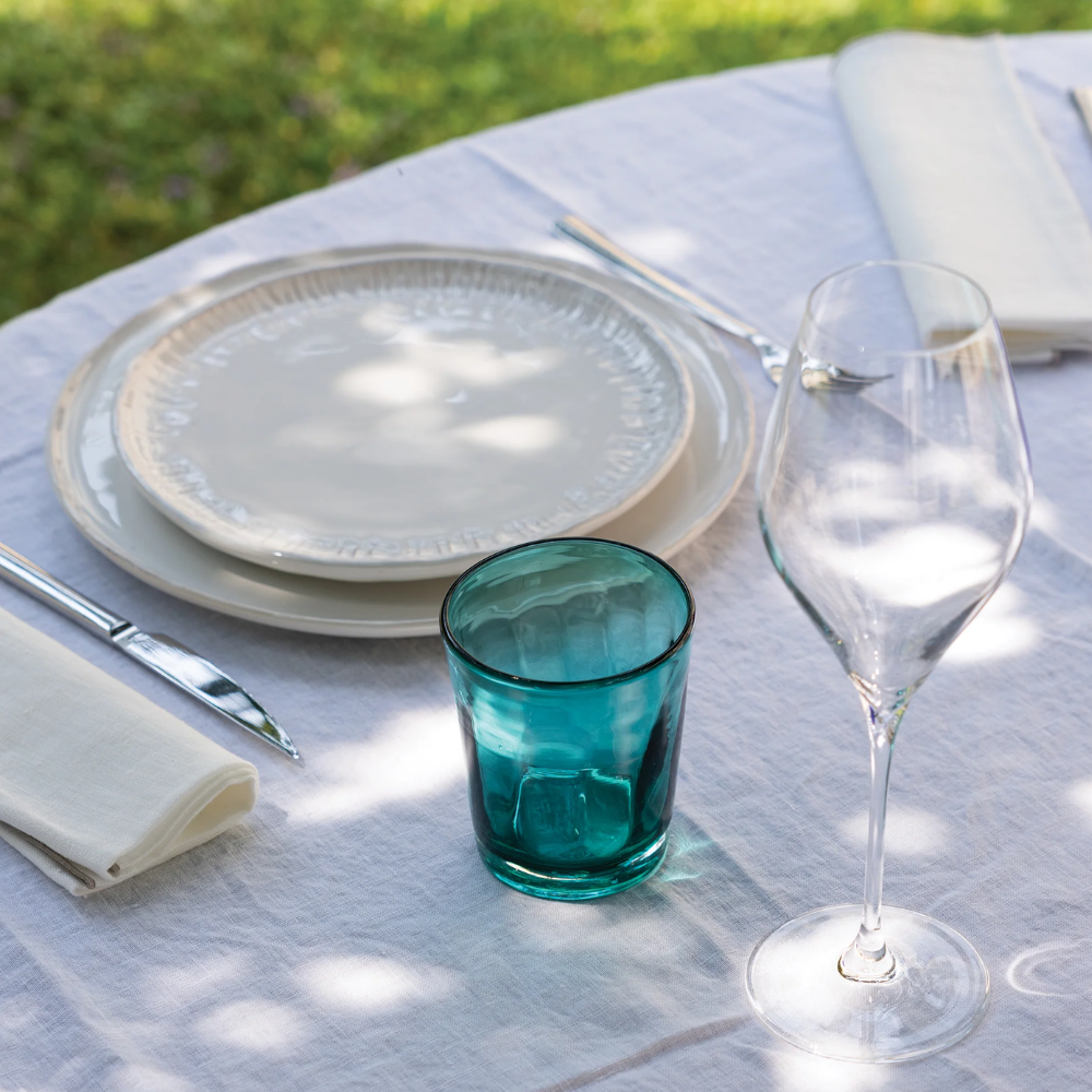 Transparent light blue glass tumbler, white plates, cutlery and wine glass on outdoor table with white tablecloth.