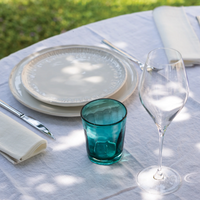 Transparent light blue glass tumbler, white plates, cutlery and wine glass on outdoor table with white tablecloth.