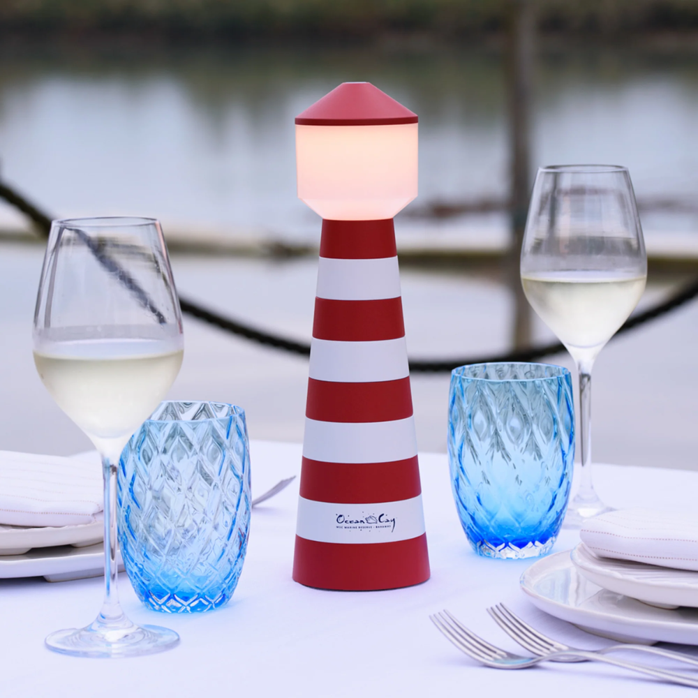 Faro decorativo rosso e bianco in legno con luce interna, due bicchieri di vino bianco, due bicchieri blu e tavola imbandita all'aperto