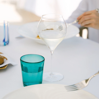 Transparent glass tumbler with acqua next to turquoise glass, white tablecloth, plates and cutlery in un restaurant setting.