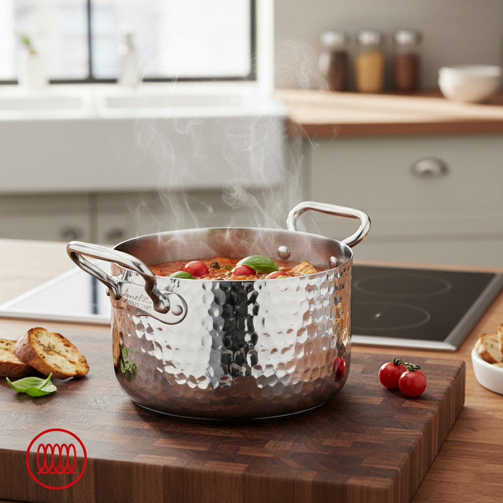 Hammered stainless steel pot with two handles full of hot sauce with cherry tomatoes and basil on wooden board in modern kitchen.