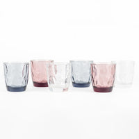 Set di sei bicchieri in vetro di diversi colori: rosa, grigio, trasparente e blu scuro, con superficie ondulata.