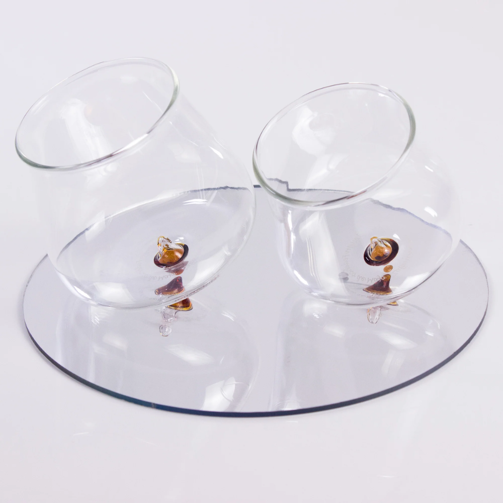 Two tilted transparent glass glasses with yellow base on round reflective mirror on white background.