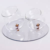 Two tilted transparent glass glasses with yellow base on round reflective mirror on white background.