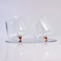 Two transparent glass glasses with amber pointed base tilted on a light grey circular reflective stand.