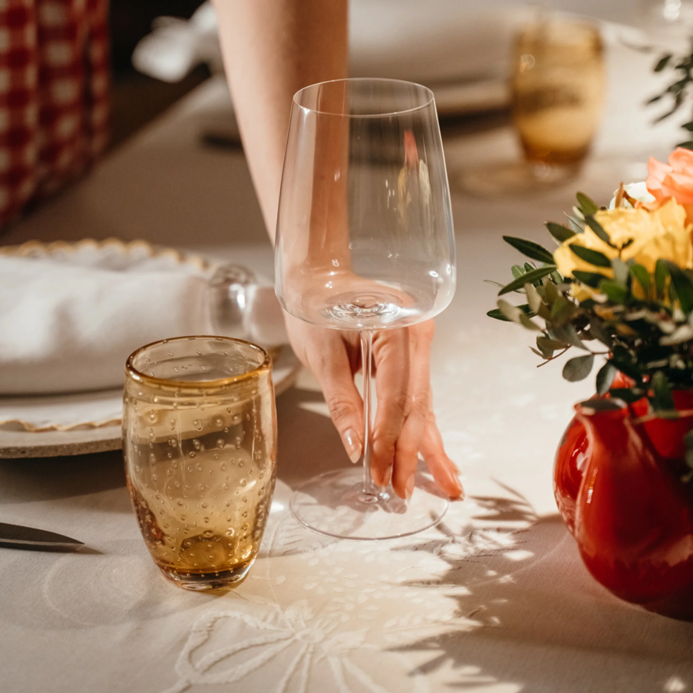 Bicchiere da vino trasparente in vetro sottile con mano che lo prende, bicchiere ambrato con bolle e vaso rosso con fiori gialli e