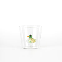 A transparent glass contains a colored duck figure with green body and yellow spots. White background.