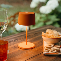 Matte orange table lamp with cylindrical shade, lit on wood table with glass and snack in bowl and plate.