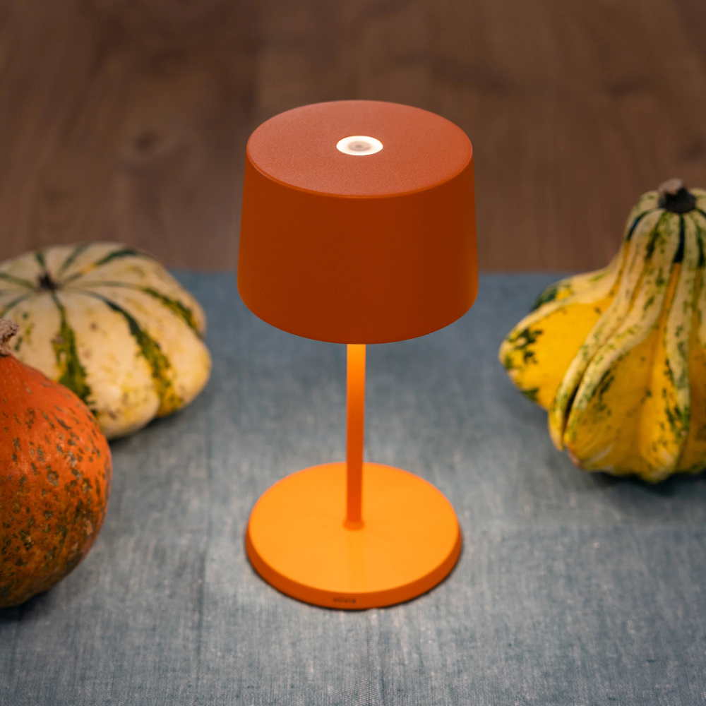 Matte orange metal table lamp with cylindrical shade and three decorative pumpkins around on blue fabric