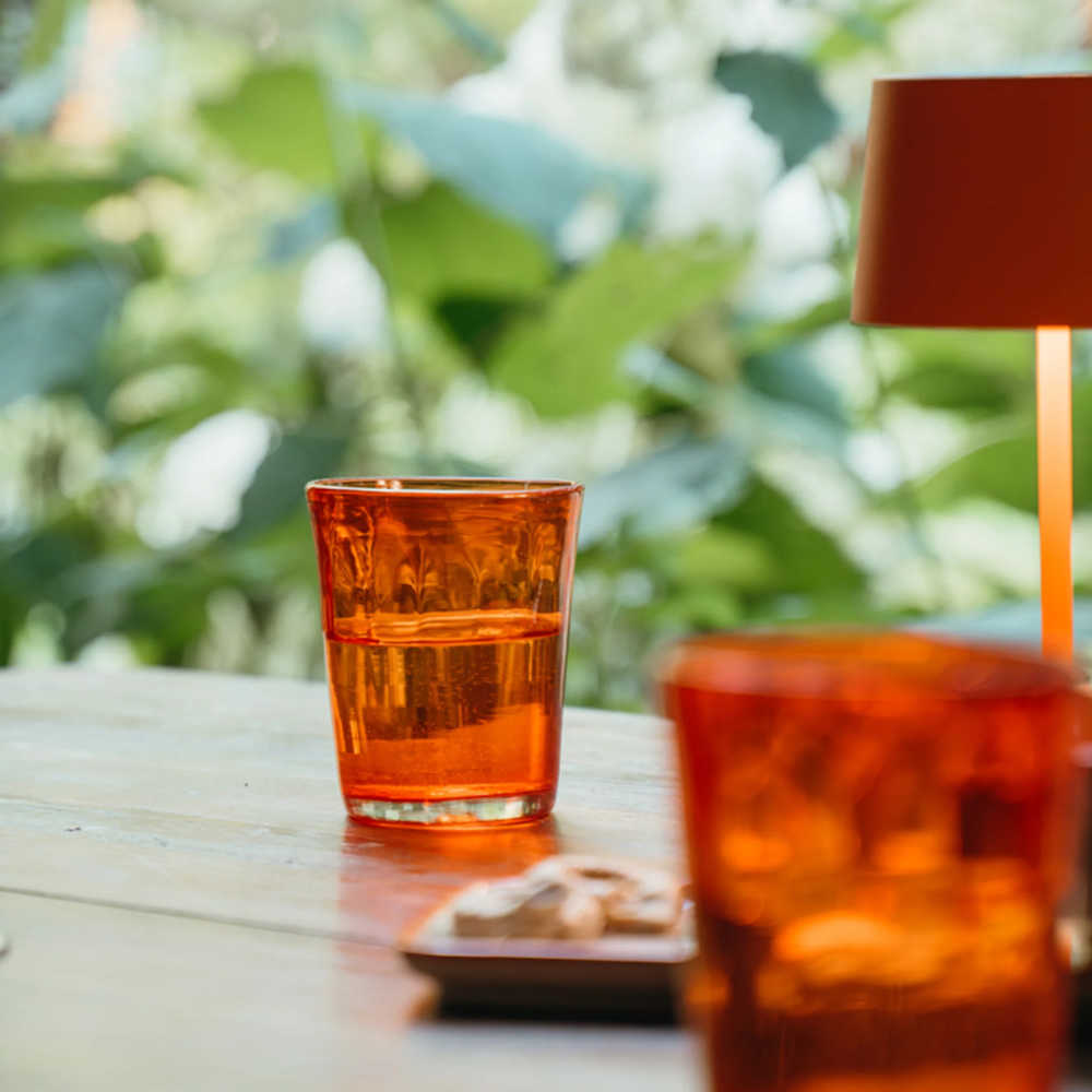 Orange glass tumbler in glass partially full on wooden table with second glass, small plate with cookies and lamp orange