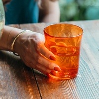 Female hand with gold bracelet and orange nails holding orange glass on wooden table.