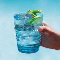 Bicchiere blu trasparente con ghiaccio, fetta di lime e foglie di menta, tenuto da una mano su sfondo azzurro sfocato.