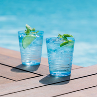 Due bicchieri azzurrati di vetro con ghiaccio, fettine di lime e foglie di menta su bordo piscina in legno chiaro, acqua azzurra sullo
