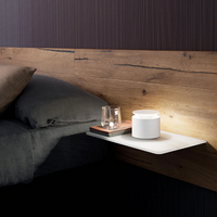 White floating nightstand with lit white cylindrical lamp, clear glass, books and light wood headboard, bed with grey blanket.