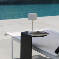 White metal table lamp on black side table next to white lounger with pool in the background.