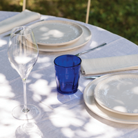 Round blue glass tumbler, white plates with decorated rim, beige napkin, cutlery and transparent wine glass on white tablecloth