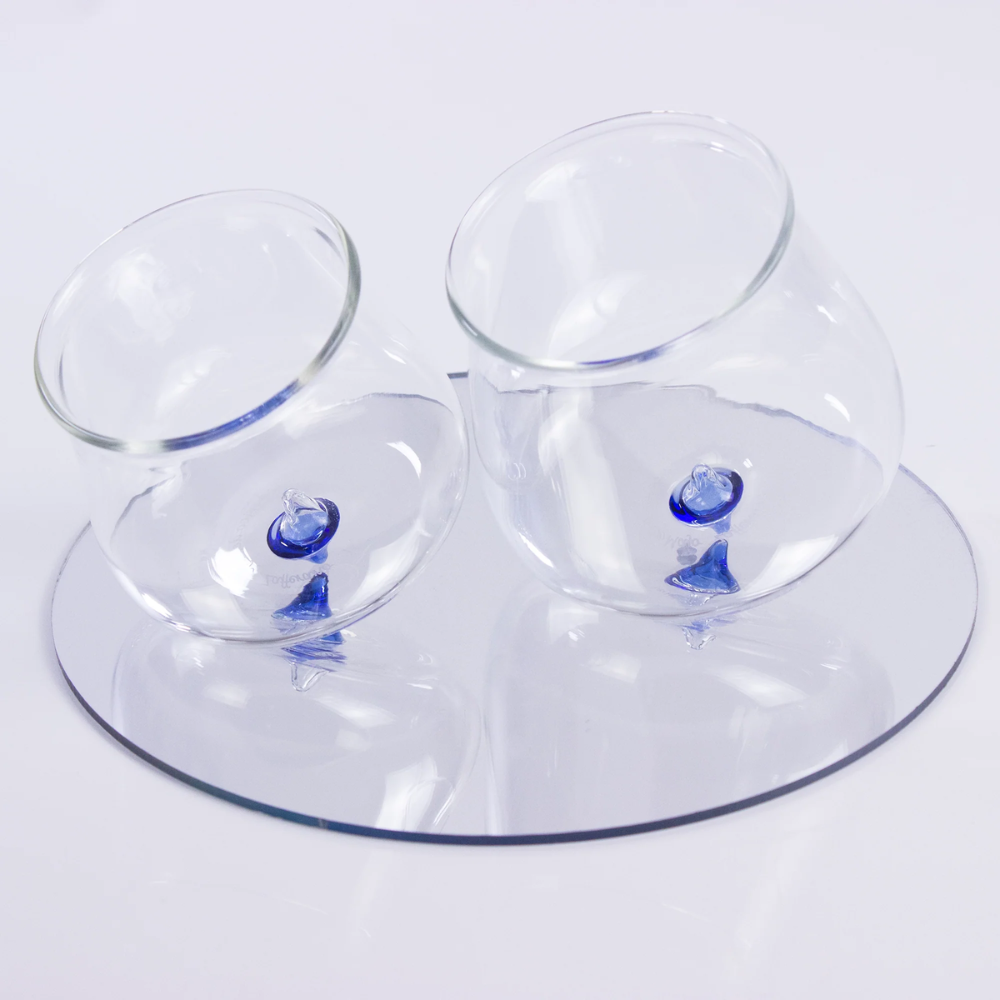 Two transparent glass glasses with round blue base resting on a white reflective transparent plate.