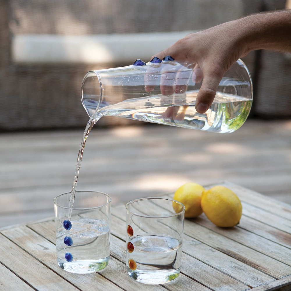 Brocca et verres transparents en verre avec décorations colorées, eau versée dans un verre sur table en bois avec citrons jaunes.
