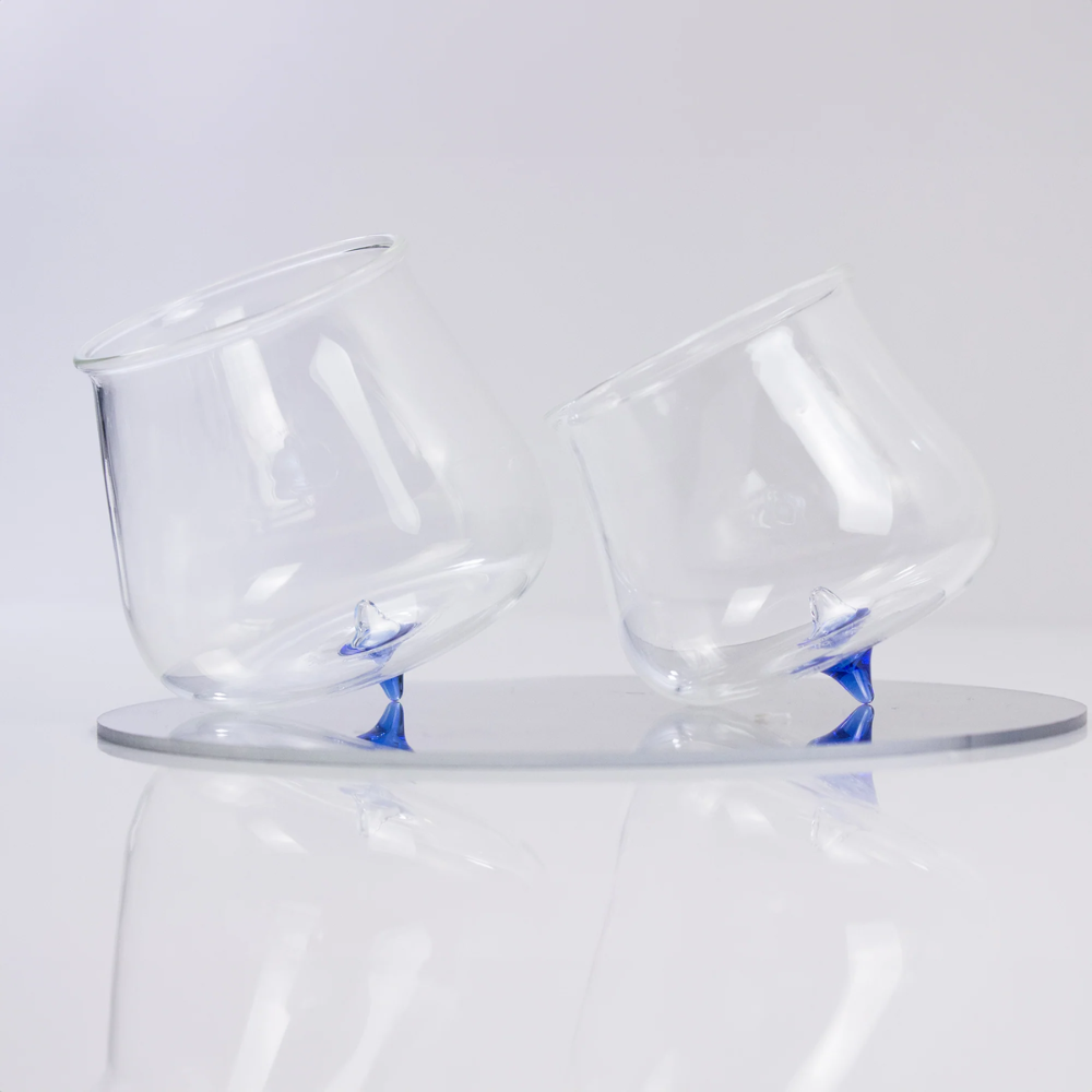 Two tilted transparent glasses with blue pointed base, resting on a light round reflective surface.