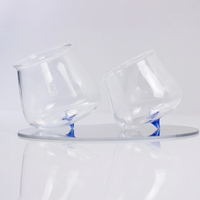 Two tilted transparent glasses with blue pointed base, resting on a light round reflective surface.