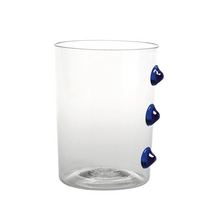 Verre transparent cylindrique avec trois décorations bleues saillantes sur un côté, fond blanc uniforme