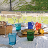 Colored glass tumblers and transparent wine glasses on outdoor table with bread on board and white tablecloth.