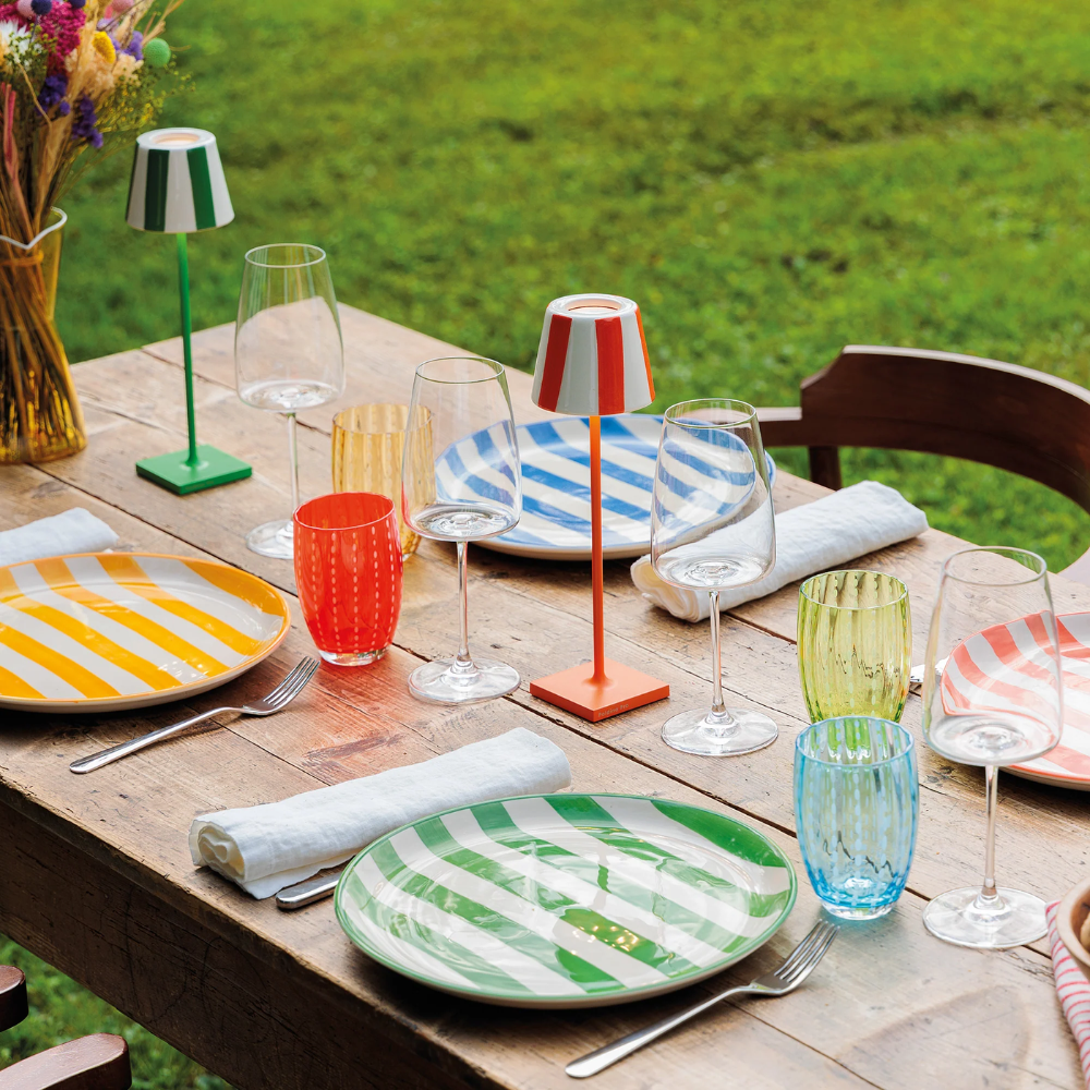 Outdoor table with striped colorful ceramic plates, transparent and colored glass glasses, cutlery and colored metal lanterns.