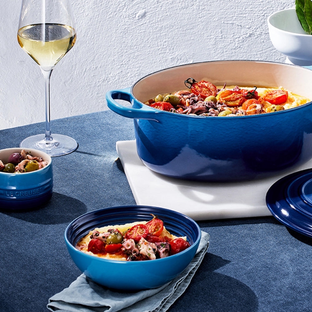 Cocotte ovale en céramique bleue avec anses, remplie de polenta, tomates cerises, olives et poulpe, accompagnée d'un verre de vin blanc et assiettes assorties