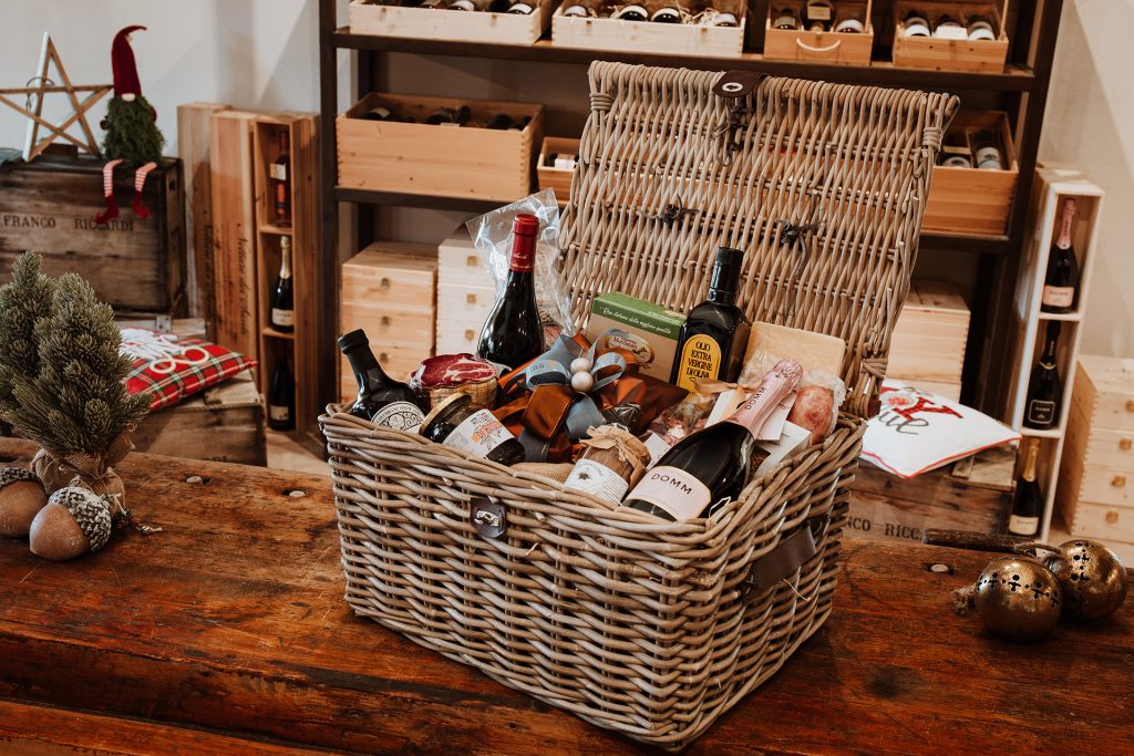 Natural wicker basket containing wine bottles, oil, cured meats and cheeses, on wooden table in a shop