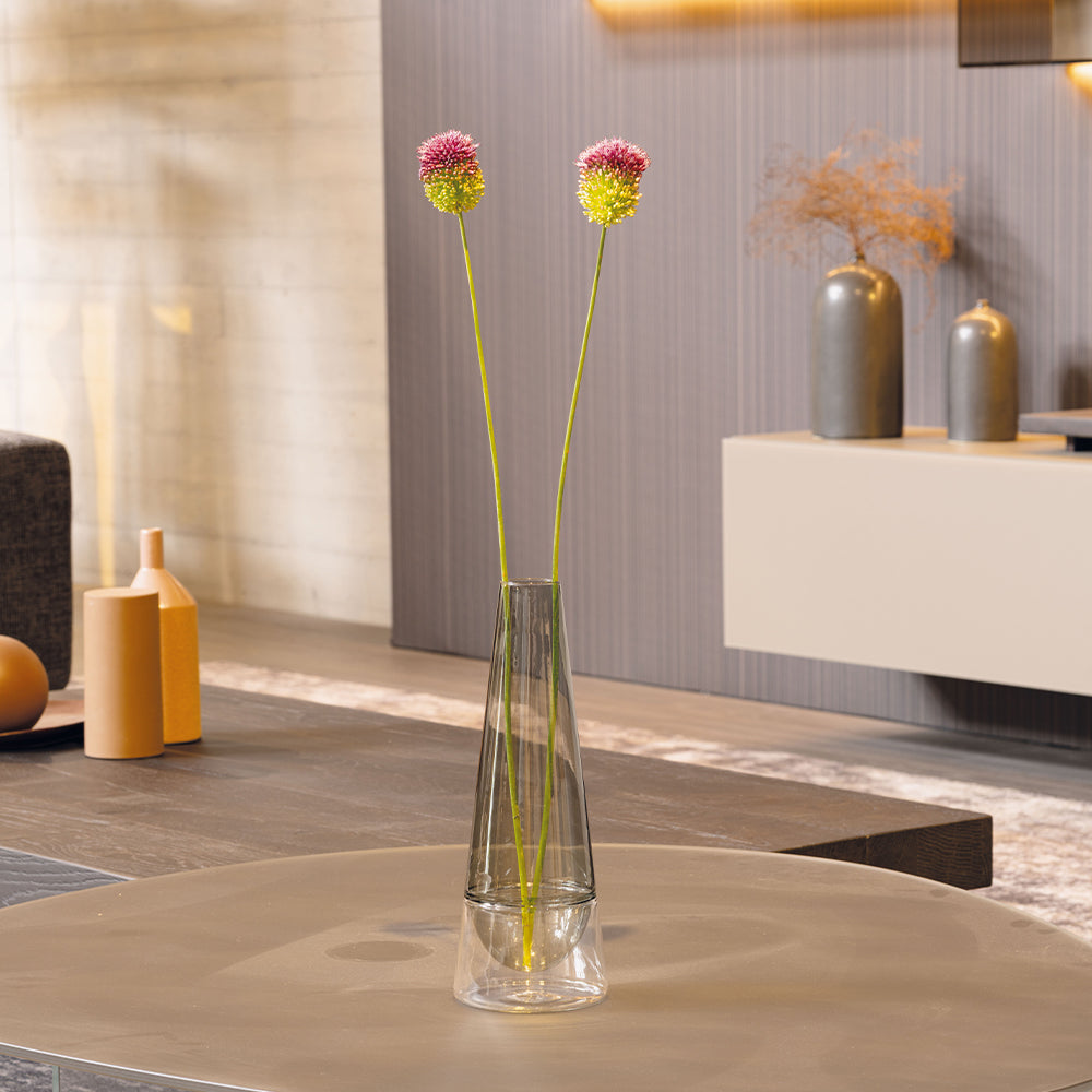 Vase transparent avec deux fleurs jaune et rose, sur table en bois, entouré d'objets décoratifs et d'un mur gris.