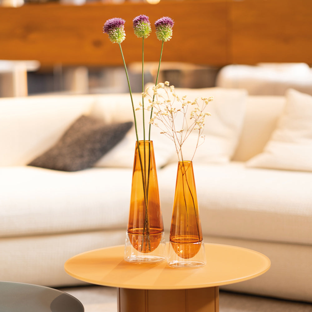 Dos jarrones de vidrio naranja en una mesita amarilla, con flores moradas y ramas secas, en un ambiente luminoso y moderno.