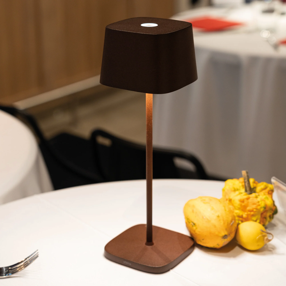 Dark brown table lamp with square base and trapezoidal shade, on white table with yellow pumpkins nearby.